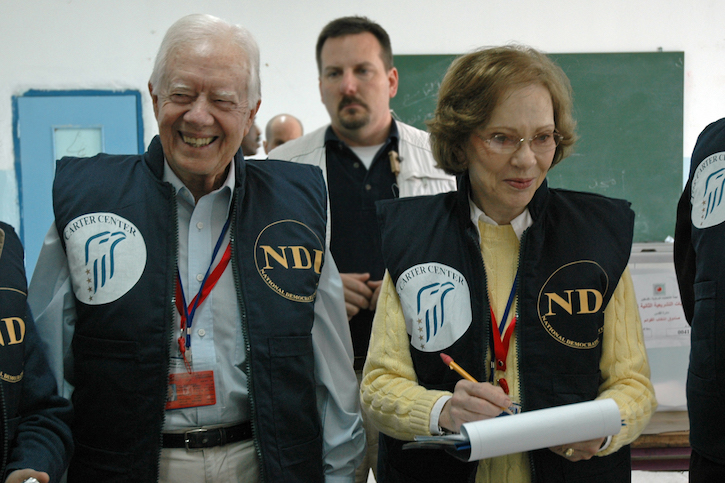 An older man and an older women in vests and professional clothing smile as they observe a polling place.
